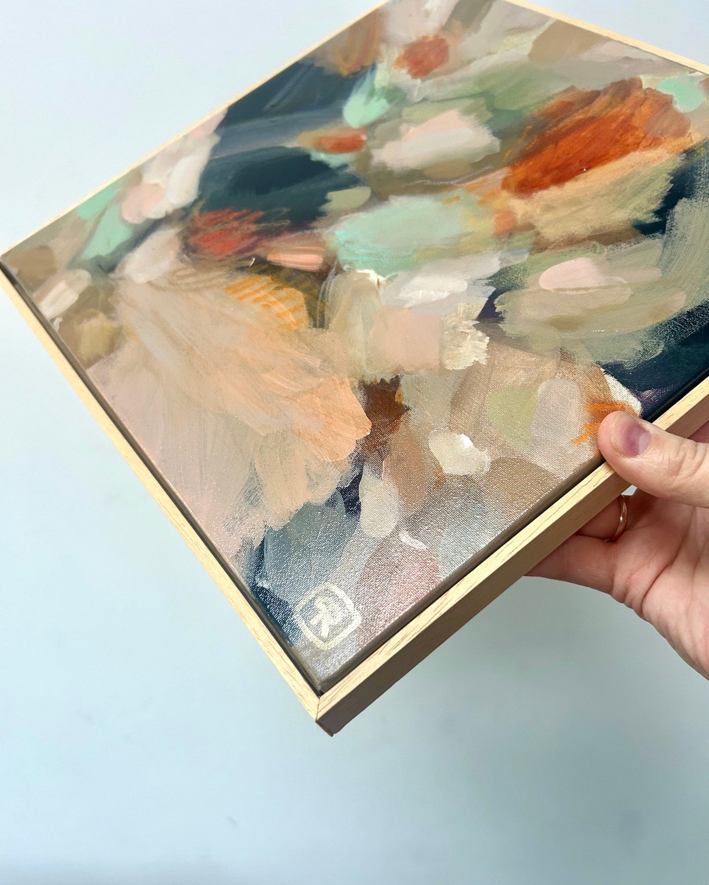 Hand holding abstract earthy tones original painting in a solid wood frame against a light background by Ruta Puidokaite.